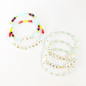 May include: A collection of six beaded bracelets featuring light blue, red, yellow, and dark blue beads with gold accents. Two bracelets display the text TS5 and TAY STORY, while four bracelets feature the repeating text I KNEW YOU KNEW IT.