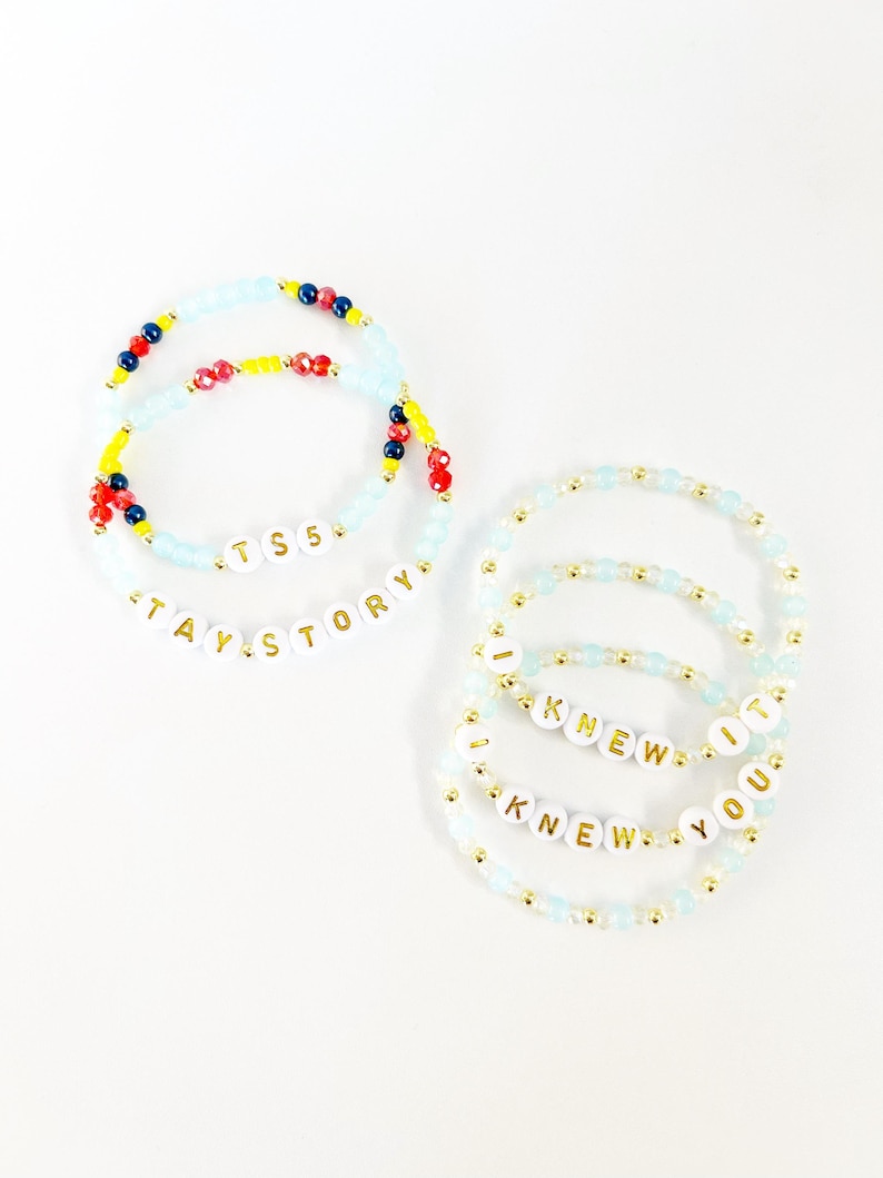May include: A collection of six beaded bracelets featuring light blue, red, yellow, and dark blue beads with gold accents. Two bracelets display the text TS5 and TAY STORY, while four bracelets feature the repeating text I KNEW YOU KNEW IT.