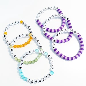 May include: A collection of beaded bracelets with white letter beads spelling out phrases like "PLEASE LEAVE", "GARBAGE IS STILL GARBAGE", "FRIENDS 4 EVER", and "JUST MARRIED". The bracelets feature various colors, including orange, green, blue, and purple.