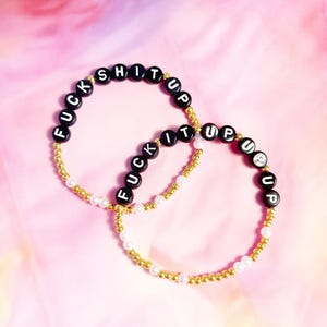 May include: Two beaded bracelets with the words "FUCK SHIT UP" spelled out in black letter beads. The bracelets have gold and white beads. The background is a pink and white gradient.