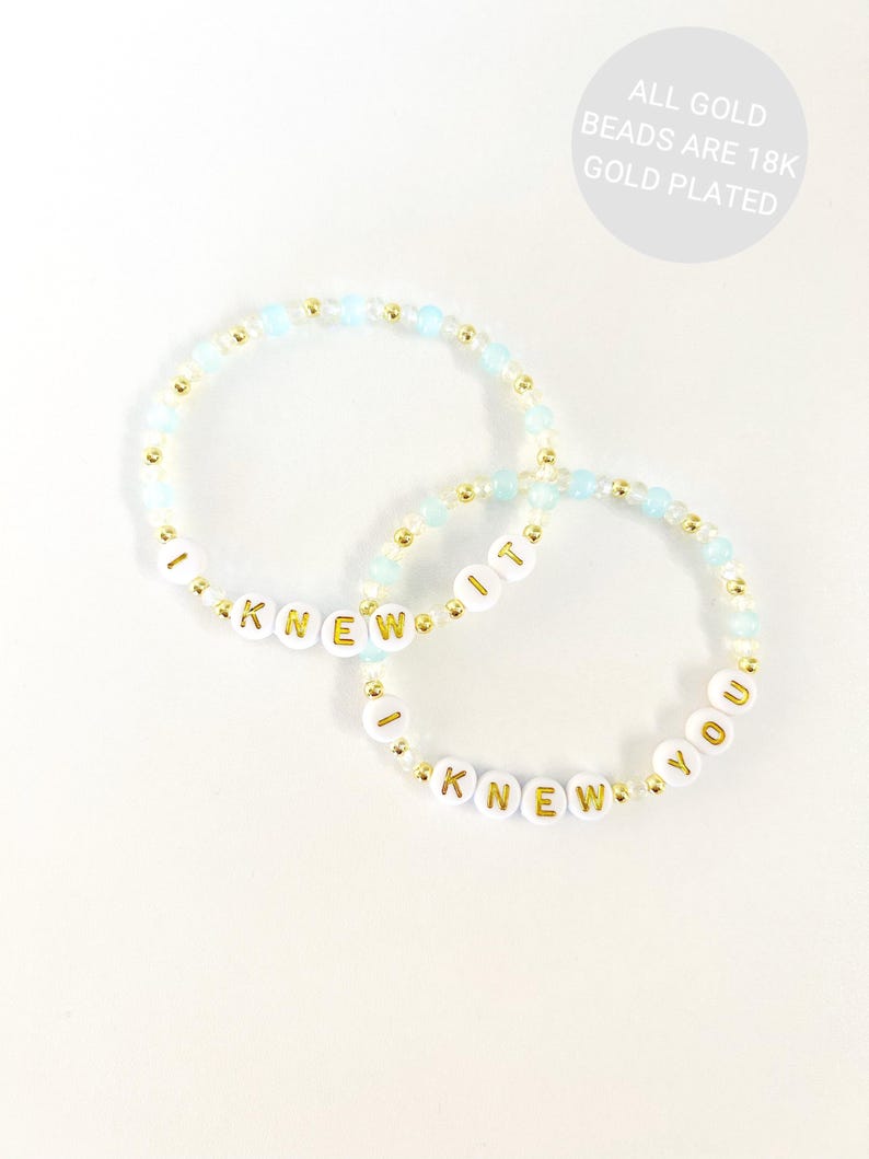 May include: Two stretch bracelets featuring light blue and white beads, small gold-tone beads, and white letter beads with gold text. One bracelet spells I KNEW IT and the other spells I KNEW YOU. Text on image: ALL GOLD BEADS ARE 18K GOLD PLATED.