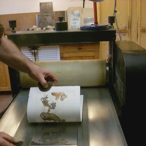 May include: A printing press in operation, with a person using a hand tool to apply pressure to a printed image. The image features brown and tan illustrations on white paper. The press is black and silver.