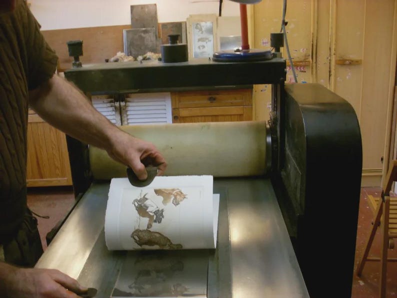 May include: A printing press in operation, with a person using a hand tool to apply pressure to a printed image. The image features brown and tan illustrations on white paper. The press is black and silver.