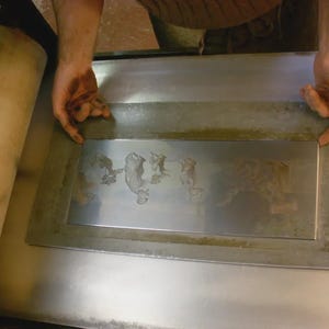 May include: A metal printing plate with an etched design, held by hands. The plate is rectangular with a raised border and shows an image of figures. A large, cylindrical roller is visible on the left.
