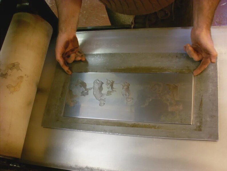 May include: A metal printing plate with an etched design, held by hands. The plate is rectangular with a raised border and shows an image of figures. A large, cylindrical roller is visible on the left.