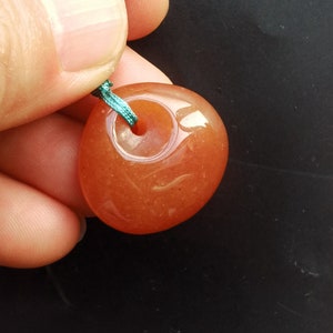 Może przedstawiać: Gładki, okrągły, pomarańczowy wisiorek z karneolu z otworem na sznurek. Wisiorek jest zawieszony na sznurku turkusowym.