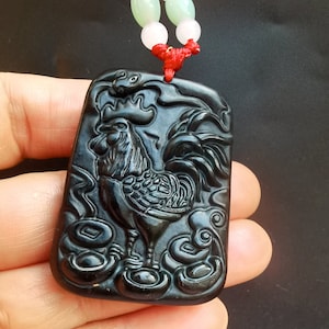 Puede incluir: Péndulo de jade negro tallado con un gallo y monedas chinas. El colgante está unido a una cuerda roja con cuentas blancas y verdes.