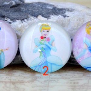 May include: Three oval-shaped glass cabochons with images of Disney princesses. The first cabochon features Jasmine from Aladdin in a blue and gold outfit. The second cabochon features Cinderella in a blue gown holding a flower. The third cabochon features Cinderella in a blue gown with a pink background.