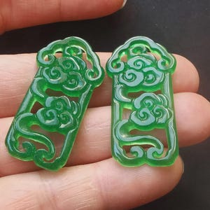 Pode incluir: Dois pingentes de jade verde translúcido. Cada pingente tem formato retangular com um desenho de nuvem esculpido na superfície. O jade tem um acabamento brilhante. Os pingentes são segurados pelos dedos de uma pessoa.
