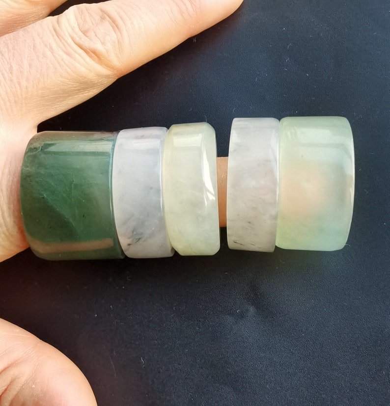 May include: Five jade rings stacked on a finger. The rings are different sizes and shades of green and white.
