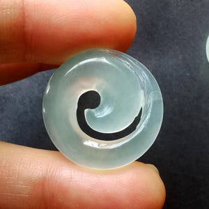 Pode incluir: Um pingente redondo de jade verde pálido com um design espiral esculpido. O pingente é segurado entre dois dedos contra um fundo escuro. A espiral é de uma cor escura e contrastante.