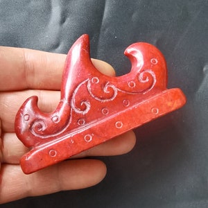 May include: A red jade carving of a stylized wave design. The carving is detailed with intricate swirls and dots.