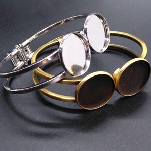 May include: Two silver and gold bangle bracelets with round, clear glass cabochons. The cabochons are set in the bracelets and are ready to be filled with resin or other materials.