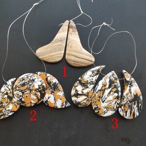 May include: Three pairs of gemstone pendants, each pair with a unique pattern. The first pair is a light brown colour with a swirling pattern. The second pair is a black, white, and orange colour with a marbled pattern. The third pair is a black, white, and orange colour with a marbled pattern.