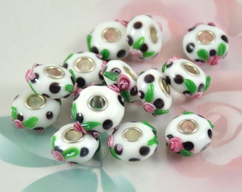 May include: A group of white glass beads with black polka dots and pink and green floral accents. The beads have a hole in the centre for stringing.