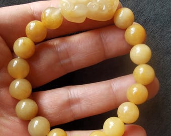 Op de afbeelding: Een goudgele kralenarmband met een gesneden middelpunt. De armband heeft gladde, ronde kralen en een gedetailleerd, gesneden middelpunt. De kralen en het middelpunt hebben een gepolijste, glanzende afwerking. De armband is een sieraad.