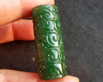 May include: A cylindrical, dark green jade pendant with intricate carved scrollwork. The jade is polished, reflecting light and highlighting the detailed design. The pendant is held between fingers.