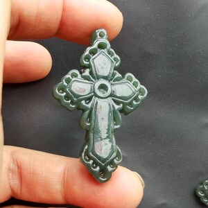 May include: A green jade cross pendant with intricate detailing. The cross is carved with a detailed design and has a smooth, polished finish.