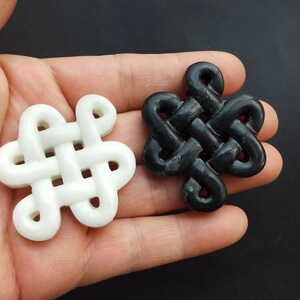 Op de afbeelding: Twee ingewikkeld gesneden knoop hangers, een wit en een zwart, gemaakt van steen. De knopen zijn een traditioneel symbool van geluk en fortuin.