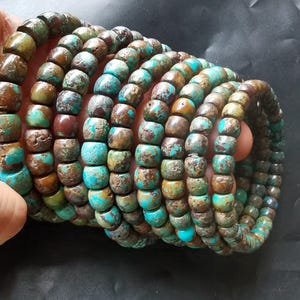 Peut inclure: Plusieurs bracelets de perles aux tons turquoise, marron et vert. Les perles cylindriques sont faites d'une pierre naturelle. Les bracelets sont empilés, mettant en valeur les variations de couleurs et les textures des perles.