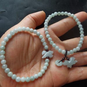 Peut inclure: Deux bracelets en jade avec des breloques d'oiseaux sculptés. Les bracelets sont faits de perles de jade vert clair et ont un cordon brun foncé.