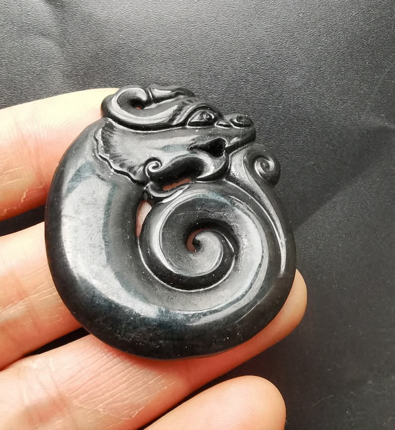 May include: A black jade pendant carved in the shape of a dragon with a swirling tail. The pendant is smooth and polished.