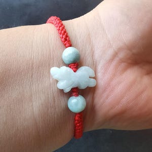 May include: Red braided bracelet with a light green jade animal carving, possibly a dog or horse. The bracelet also has two light green jade beads. The bracelet is worn on a wrist.