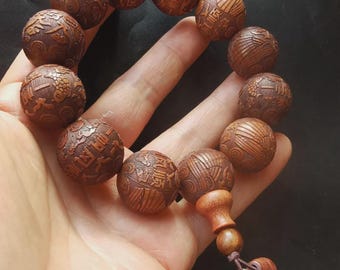 May include: A wooden bracelet with intricately carved spherical beads. The beads are a warm brown color and feature detailed patterns and Chinese characters. The bracelet is held in a hand against a dark background, showcasing the craftsmanship.