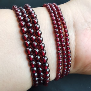May include: Four strands of dark red garnet bead bracelets stacked on a person's wrist.