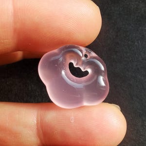 May include: A translucent, pink gemstone pendant in the shape of a cloud with a heart-shaped cutout. The pendant has a small hole at the top for stringing. The gemstone is held between two fingers against a dark background.