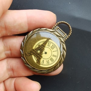 Puede incluir: Un reloj de bolsillo de color bronce con una cara de vidrio que presenta una silueta de la Torre Eiffel y una esfera de reloj con números romanos.