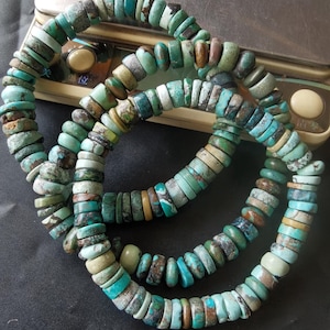 May include: A strand of disc-shaped turquoise beads featuring natural variations in colour, ranging from light sky blue to deep teal and earthy brown matrix patterns. The beads are strung together in a continuous loop, displaying a smooth, polished finish.