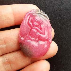 May include: A pink and gray carved stone pendant featuring a monkey design. The pendant is oval shaped and has a smooth, polished finish.