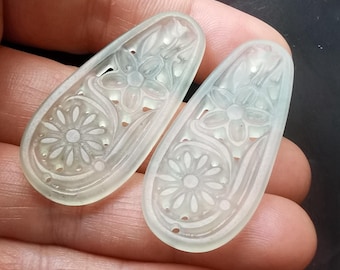 May include: Two pale green, translucent, teardrop-shaped pendants featuring intricate, carved floral motifs. Each piece displays a central flower design with radiating petals and curved stems, finished with a smooth, polished surface texture.