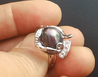 May include: A silver ring with a large iridescent black pearl held in a stylized hand design. The ring is adorned with small clear cubic zirconia stones.