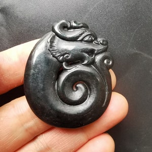 May include: A black jade pendant carved in the shape of a dragon. The dragon is curled up in a spiral shape, with its head facing to the right. The pendant is smooth and polished.