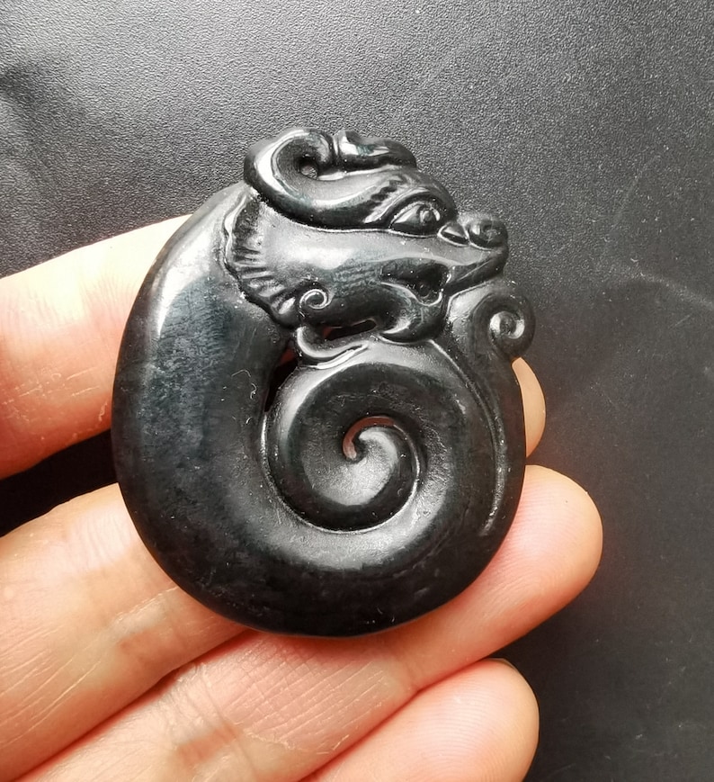 May include: A black jade pendant carved in the shape of a dragon. The dragon is curled up in a spiral shape, with its head facing to the right. The pendant is smooth and polished.