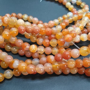 May include: A strand of orange carnelian beads. The beads are round and smooth, with a translucent finish. The beads are strung on a clear elastic cord.