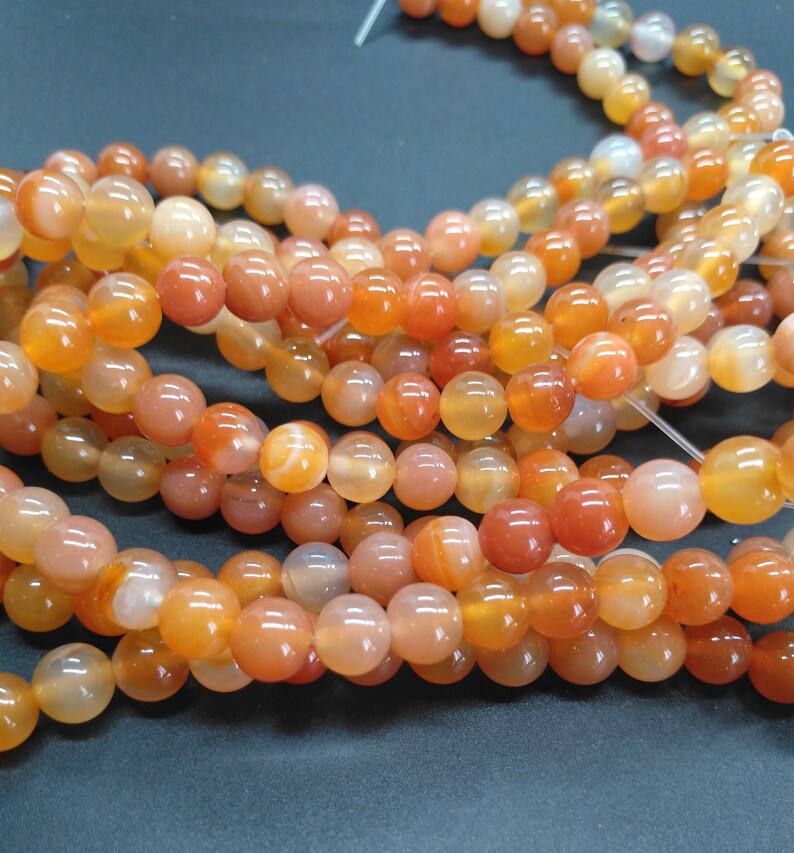 May include: A strand of orange carnelian beads. The beads are round and smooth, with a translucent finish. The beads are strung on a clear elastic cord.