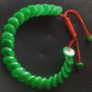 May include: A jade bracelet featuring a series of bright green, circular beads. The beads are strung together with a red cord that forms the adjustable closure. The bracelet is designed to be worn around the wrist.