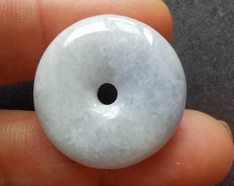 May include: A round, pale blue-green jade pendant with a central hole. The polished stone has a translucent quality, with subtle variations in color. The pendant is held between fingers, showcasing its smooth surface and natural beauty.