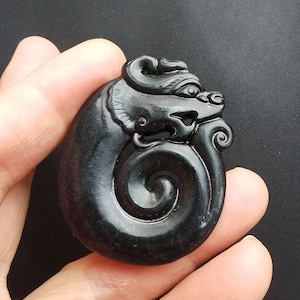 May include: A black jade pendant carved in the shape of a dragon. The dragon is curled up in a spiral shape, with its head facing to the right. The pendant is smooth and polished.
