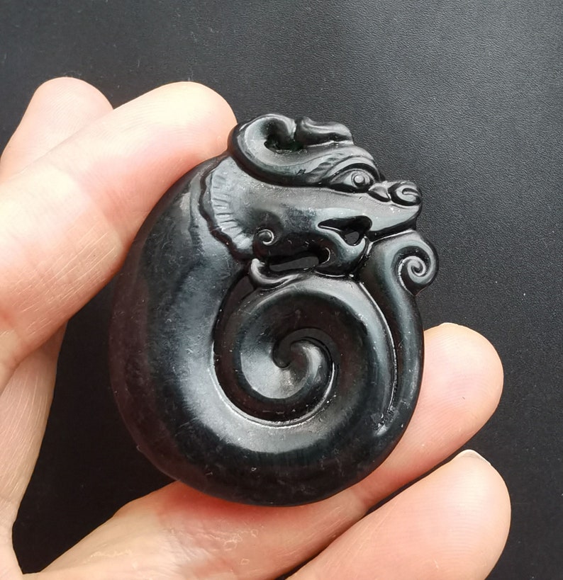 May include: A black jade pendant carved in the shape of a dragon. The dragon is curled up in a spiral shape, with its head facing to the right. The pendant is smooth and polished.