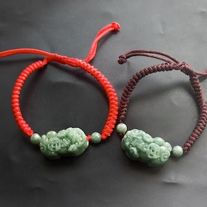 Op de afbeelding: Twee jade armbanden met groene jade bedels en rode en bruine koorden. De bedels zijn gegraveerd met een Chinees geluksymbool.