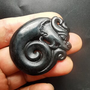 May include: A black jade pendant carved in the shape of a dragon with a curled tail. The dragon's face is detailed with eyes, a nose, and a mouth.