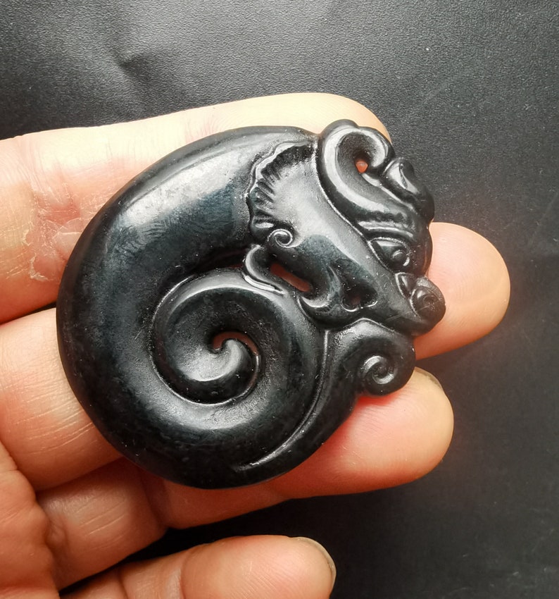 May include: A black jade pendant carved in the shape of a dragon with a curled tail. The dragon's face is detailed with eyes, a nose, and a mouth.