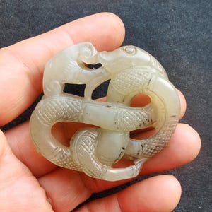 May include: A pale green jade pendant carved in the shape of two intertwined mythical creatures. The carving features detailed scales and swirling designs, with a smooth, polished finish. The pendant is held in a hand.