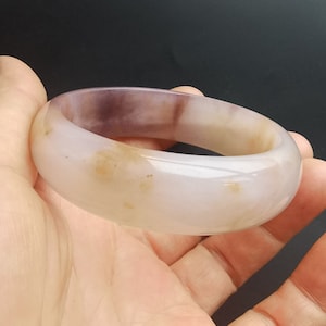 Può includere: Un braccialetto a cerchio in giada lucidata e liscia. Il braccialetto presenta una miscela di colori, tra cui rosa pallido, crema e un tocco di viola. La pietra ha una qualità traslucida, con sottili variazioni di colore e consistenza.