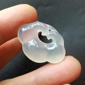 May include: A translucent, cloud-shaped pendant with a smooth, polished surface. The pendant has a curved cutout and a small hole near the top. The color is a soft, milky white, and it is held between two fingers against a dark background.