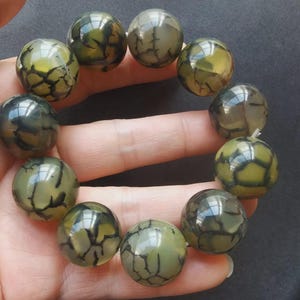 Puede incluir: Una pulsera con cuentas grandes y redondas con un patrón agrietado único. Las cuentas son principalmente de color verde oliva con vetas grises oscuras, creando un contraste llamativo. La pulsera se sostiene en una mano.